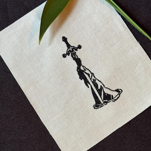 Op de afbeelding: Een vierkant stuk gebroken wit textiel met een zwarte inktillustratie van een zwaard gewikkeld in vloeiende stof. Het ontwerp is gecentreerd op het textiel, dat een zichtbare geweven structuur heeft. Een groen blad rust op de bovenrand.