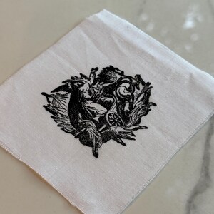 May include: White square handkerchief with a detailed black illustration. The illustration depicts a classical scene with figures and ornate details. The fabric has a subtle texture.