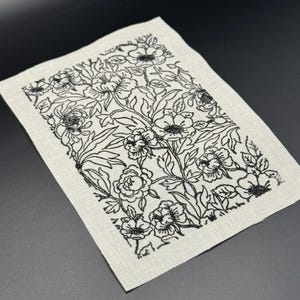 Puede incluir: Un cuadrado de tela blanca con un dise&ntilde;o floral bordado en negro. El dise&ntilde;o presenta una variedad de flores y hojas.