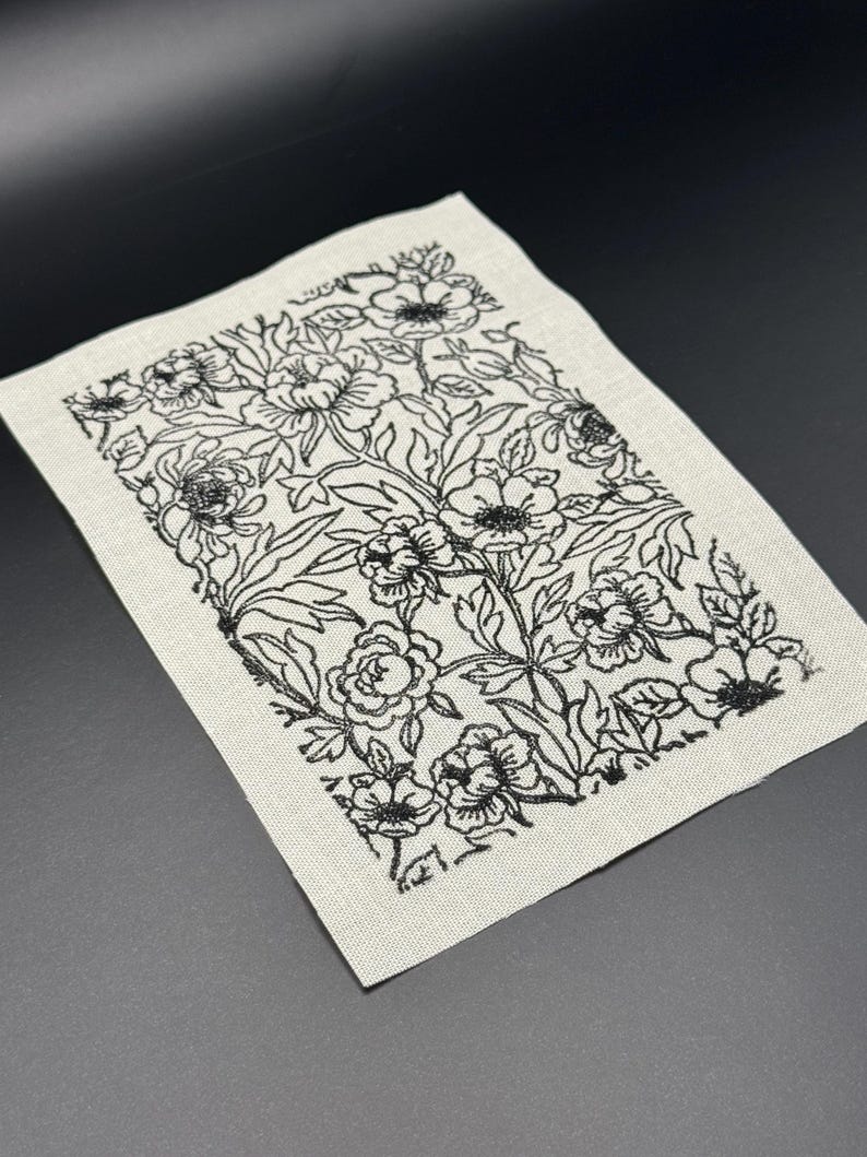 Puede incluir: Un cuadrado de tela blanca con un dise&ntilde;o floral bordado en negro. El dise&ntilde;o presenta una variedad de flores y hojas.