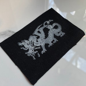 May include: A black fabric patch with a silver embroidered design of a dragon.