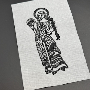 Op de afbeelding: Zwart-witte geborduurde patch met een vrouw die een staf en een bol vasthoudt. De vrouw draagt een lange jurk met een geometrisch patroon en heeft een halo boven haar hoofd.
