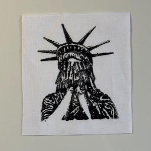 May include: A black and white patch featuring a distressed illustration of the Statue of Liberty. The statue's face is obscured, with hands clasped in front. The patch is square and made of white fabric.