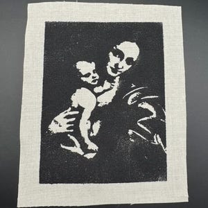 May include: Black and white image of a mother holding a baby. The image is printed on a white fabric patch.