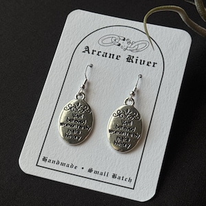 May include: A pair of silver-toned earrings with the text "Well behaved women rarely make history" on an oval pendant. The earrings hang from a white card with the text "Arcane River" and "Handmade • Small Batch".