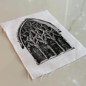 Pode incluir: Um pequeno quadrado de tecido branco com um desenho de janela gótica preta impresso. A janela apresenta intrincados padrões florais e geométricos dentro de sua moldura arqueada. As bordas do tecido são inacabadas.