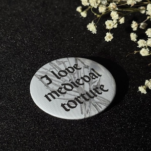 Könnte beinhalten: Ein runder, weißer Button mit dem Schriftzug "I love medieval torture" in schwarzer gotischer Schrift. Der Hintergrund zeigt eine graue Illustration mittelalterlicher Folterwerkzeuge. Der Button befindet sich auf einem schwarzen Hintergrund.