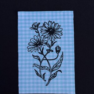 Puede incluir: Un parche de tela rectangular con un patrón de cuadros vichy en azul claro y blanco. Centrado en el parche hay una ilustración en tinta negra de dos flores tipo margarita con tallos, hojas y un capullo cerrado que brota desde la base.