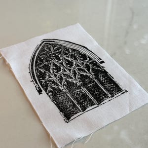 May include: A small, white fabric square featuring a black, gothic-style window design. The window has an arched top and intricate floral-like patterns within the panes. The edges of the fabric are unfinished, with loose threads.