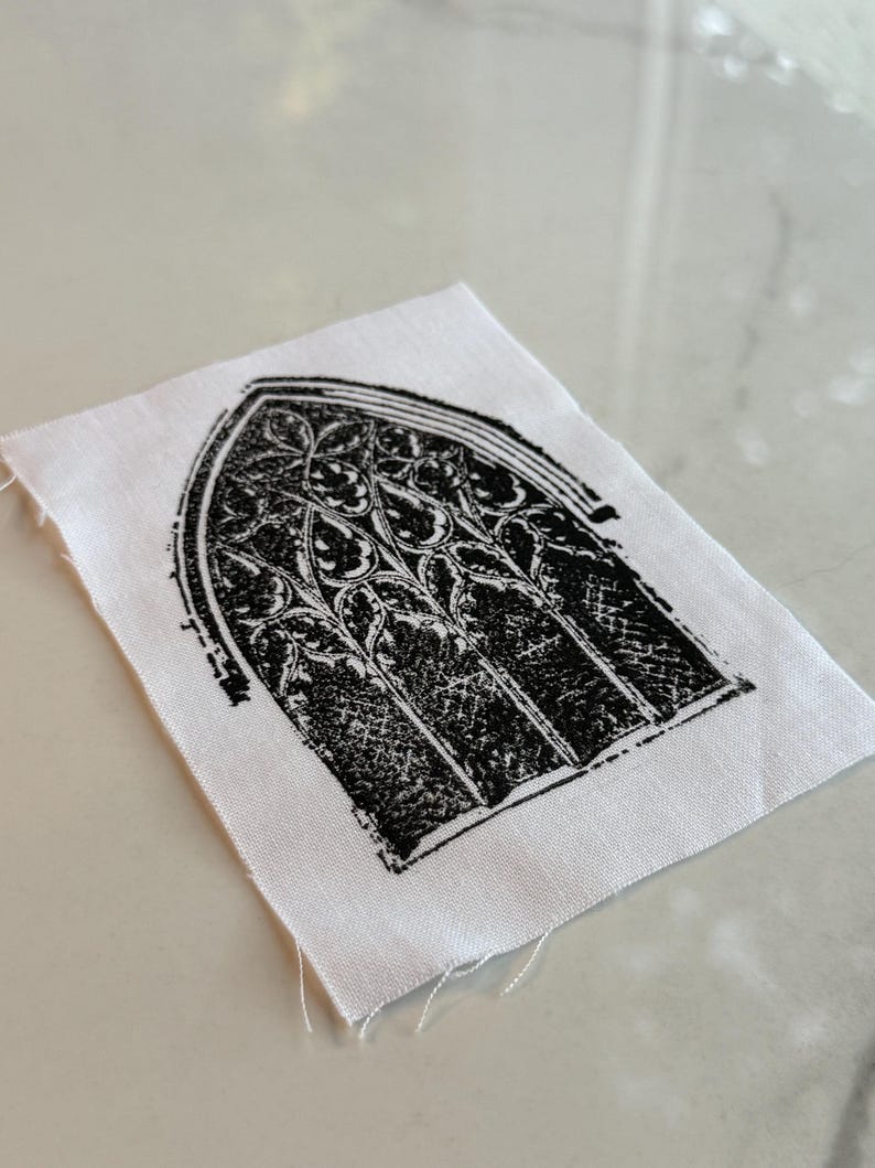 May include: A small, white fabric square featuring a black, gothic-style window design. The window has an arched top and intricate floral-like patterns within the panes. The edges of the fabric are unfinished, with loose threads.