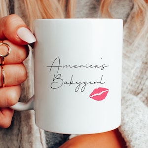 Peut inclure: Une tasse en céramique blanche avec du texte noir qui dit "America's Babygirl" et une empreinte de rouge à lèvres rose.