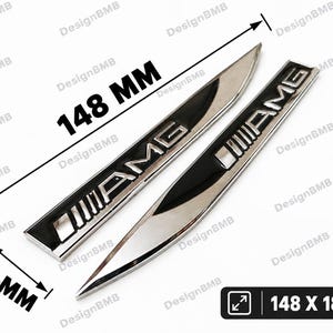 Könnte beinhalten: Zwei silberne und schwarze AMG-Autoembleme. Die Embleme zeigen den Schriftzug AMG in metallischer Ausführung. Maßlinien zeigen eine Länge von 148 mm und eine Breite von 18 mm an. Der Text 148 X 18 MM steht in einem schwarzen Kasten.