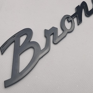 May include: A glossy black emblem featuring the word Bronco in a stylised, cursive script font. The letters are connected and have a smooth, reflective surface, showcasing a distinct, bold typography design with sharp, clean edges throughout.