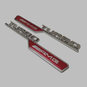 Puede incluir: Dos emblemas de coche de metal plateado con acentos rojos. Los emblemas dicen "TURBO" y "AMG".