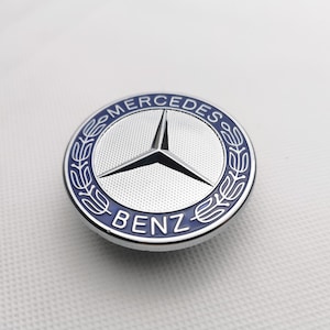 Puede incluir: Un emblema plateado de Mercedes-Benz con una corona de laurel azul y las palabras "Mercedes" y "Benz" en letras azules.