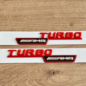 May include: Two red and silver "TURBO AMG" emblems on white backing. The word "TURBO" is in bold red letters. The "AMG" logo is in a black rectangle with silver accents. Automotive accessories.