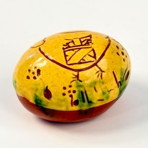 May include: A yellow ceramic egg with brown and green painted designs. The egg has a brown base and a yellow top with a brown design of a house with a chimney.