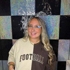 May include: A two-toned crewneck sweatshirt featuring a split design with one side in cream and the other in dark brown. The word FOOTBALL is printed in a bold, arched, dark brown font across the chest, centered over the two-toned fabric.
