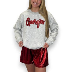 Puede incluir: Una sudadera gris claro con la palabra "Georgia" en escritura roja brillante. La sudadera se combina con pantalones cortos rojos brillantes. El atuendo es informal y adecuado para el uso diario.