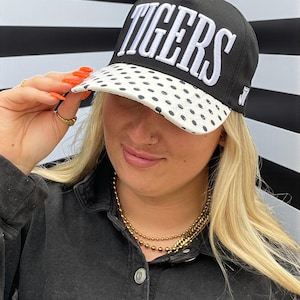 May include: A black baseball cap featuring the word TIGERS embroidered in white block letters on the front crown. The brim of the cap is white with a black polka dot pattern. A small white letter J is embroidered on the side of the cap near the brim.
