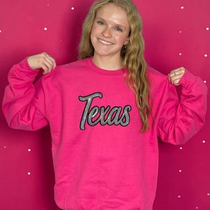 May include: A bright pink sweatshirt with the word "Texas" in a silver glitter script across the chest. The sweatshirt is a solid color and has long sleeves. The background is a pink wall with white dots.