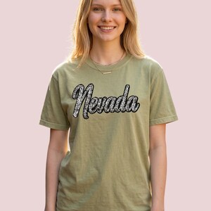 May include: Olive green t-shirt with the word "Nevada" in a cursive font with a black outline and a white and silver pattern. The person is wearing blue jeans and a gold necklace.