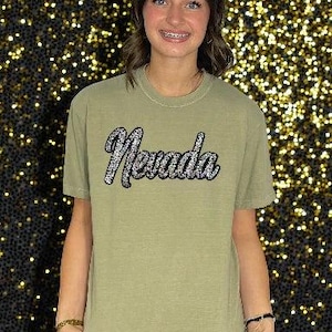 May include: An olive green t-shirt with the word "Nevada" in a sparkly, silver script across the chest. The shirt has short sleeves and a crew neck. The background is a dark color with many small, sparkling lights.