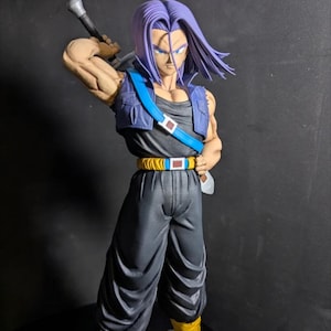 Puede incluir: Figura detallada de un personaje con pelo morado puntiagudo, que lleva una camiseta negra, un chaleco azul y pantalones grises. La figura sostiene una espada sobre su hombro. La figura tiene botas amarillas y un cinturón amarillo con una hebilla blanca.