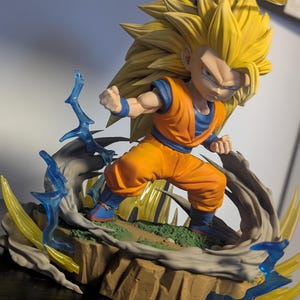 Puede incluir: Figura detallada de un personaje con pelo amarillo puntiagudo, vistiendo un atuendo naranja y azul, en una pose de lucha dinámica. La figura está sobre una base rocosa con efectos de energía azules y amarillos, y un halo sobre la cabeza.