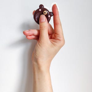 May include: A small, round, dark brown bear-shaped charm with two small ears, a single visible eye, and two light pink dots on the cheeks. The charm features the text "made by wenderdesign" in small white letters along the top edge of the object.