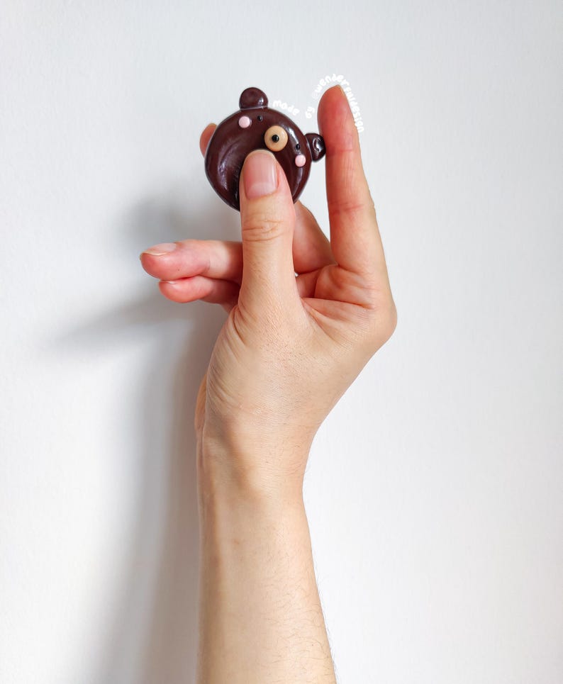 May include: A small, round, dark brown bear-shaped charm with two small ears, a single visible eye, and two light pink dots on the cheeks. The charm features the text "made by wenderdesign" in small white letters along the top edge of the object.
