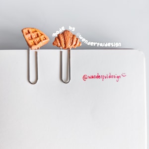 Puede incluir: Dos clips de papel metálicos con adornos en miniatura con forma de comida. Un clip presenta un gofre, el otro un cruasán. El gofre y el cruasán son de color marrón claro. También se ve el texto "made by @wonderfuldesign" y "@wonderfuldesign".