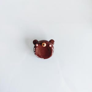 May include: A small, round, brown ceramic dish shaped like a bear face. It features two small ears, a central snout with a black nose, and two pink circular cheeks. The text "made by wonderfullesign" is printed in white along the lower left edge.