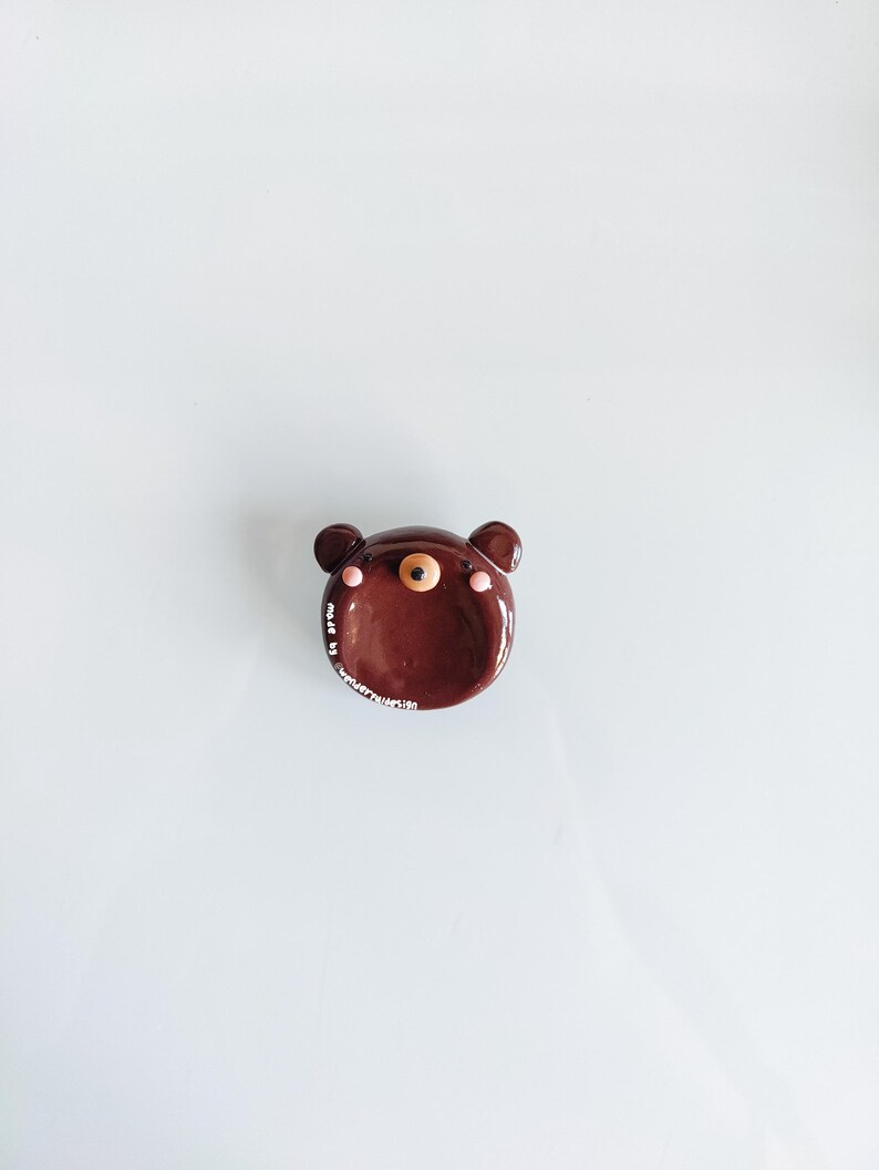May include: A small, round, brown ceramic dish shaped like a bear face. It features two small ears, a central snout with a black nose, and two pink circular cheeks. The text "made by wonderfullesign" is printed in white along the lower left edge.