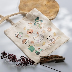 Könnte beinhalten: Eine beigefarbene Stofftasche mit einem bunten Aufdruck herbstlicher Motive wie Eulen, Kürbissen, Eicheln, einer Tasse heißer Schokolade, einem Buch und einem Zaubertrankfläschchen sowie dem Schriftzug fall vibes in schwarzer Schreibschrift.