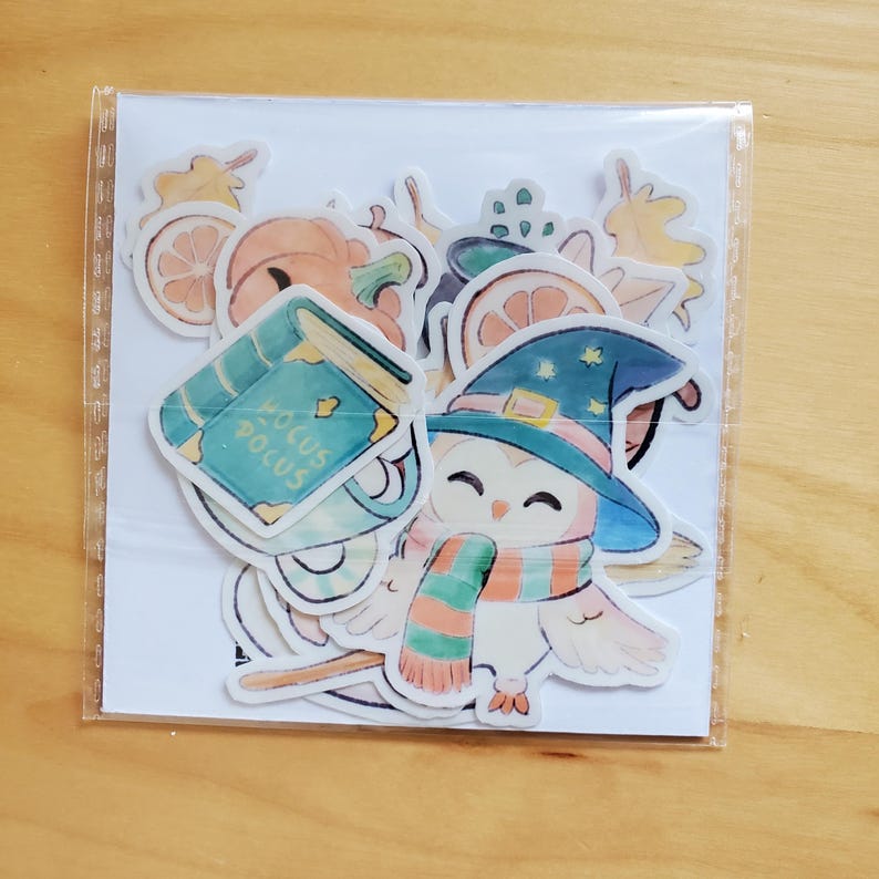 May include: A collection of colorful stickers featuring autumn and Halloween themes. Designs include a witch owl wearing a hat and scarf, a pumpkin, orange slices, and a book with the text "Hocus Pocus". The stickers are packaged in a clear plastic sleeve.