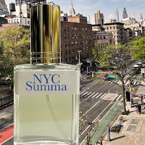 May include: A rectangular perfume bottle with a gold and silver cap. The bottle has a white label with the text "NYC Summa" in blue. The background shows a city street with buildings and trees.