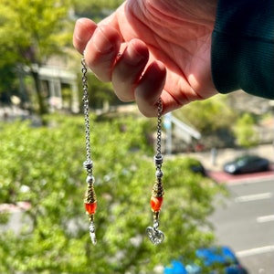 Puede incluir: Un par de pendientes de cadena plateada con un dije de corazón y una cuenta roja y dorada al final de cada cadena.