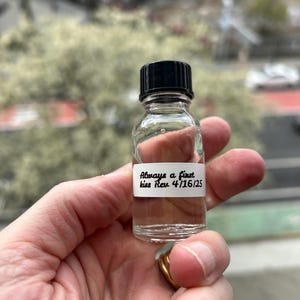 May include: A clear glass bottle with a black cap, containing a liquid. The bottle has a white label with the text "Always a first kiss Rev 4/16/25".