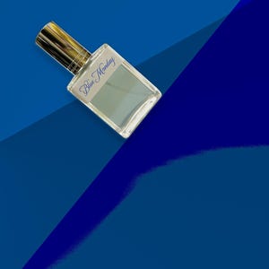 May include: A clear glass perfume bottle with a gold-colored cap and the words "Blue Monday" in blue script. The bottle is filled with a light blue liquid and is set against a backdrop of various shades of blue.