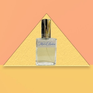 May include: A rectangular perfume bottle with a gold cap and a label that reads "Miel d'Ambre" in blue script. The bottle is filled with a light amber-colored liquid. The background is a geometric design in shades of orange and yellow.