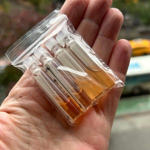 May include: A clear plastic bag containing several small glass vials filled with amber-colored liquid. The vials have white caps and are held in an open hand. The background is blurred, showing hints of greenery and a building.
