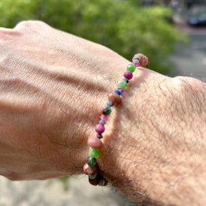 Puede incluir: Un brazalete de cuentas multicolores con cuentas marrones, verdes, rosas y azules. El brazalete está en la muñeca de una persona.