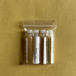 May include: A clear plastic bag containing four small glass vials with white caps. One vial has a white label with the text "The shadow within 1 Wrong". The vials are filled with a light amber liquid, set against a yellow background.