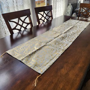 May include: A rectangular, light gray table runner with gold-colored ornate patterns and tassels at each end. The runner is placed on a dark brown wooden table, with three chairs visible in the background.