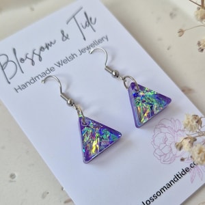 May include: A pair of handmade purple triangle earrings with iridescent accents. The earrings are displayed on a white card with the text "Blossom & Tide Handmade Welsh Jewellery" and the website address.