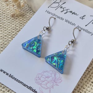 May include: A pair of handmade blue triangle earrings with iridescent accents. The earrings are suspended from silver-colored hooks. The earrings are displayed on a card with the text "Blossom & Tide Handmade Welsh Jewellery" and the website address.