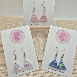May include: Three pairs of triangle-shaped earrings with iridescent glitter. The earrings are displayed on white cards with the text "Blossom & Tide" and the social media handle "@blossomandtide".