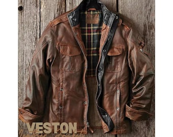 May include: A brown leather jacket featuring a black inner collar, a front zipper closure with snap buttons, two chest pockets, and a plaid lining in red, yellow, and black. The word VESTON is printed in large letters at the bottom left corner.