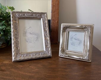 May include: Two silver picture frames with ornate designs. The larger frame is on the left and the smaller frame is on the right. Both frames have a white mat and a black and white image of a woman in a dress.